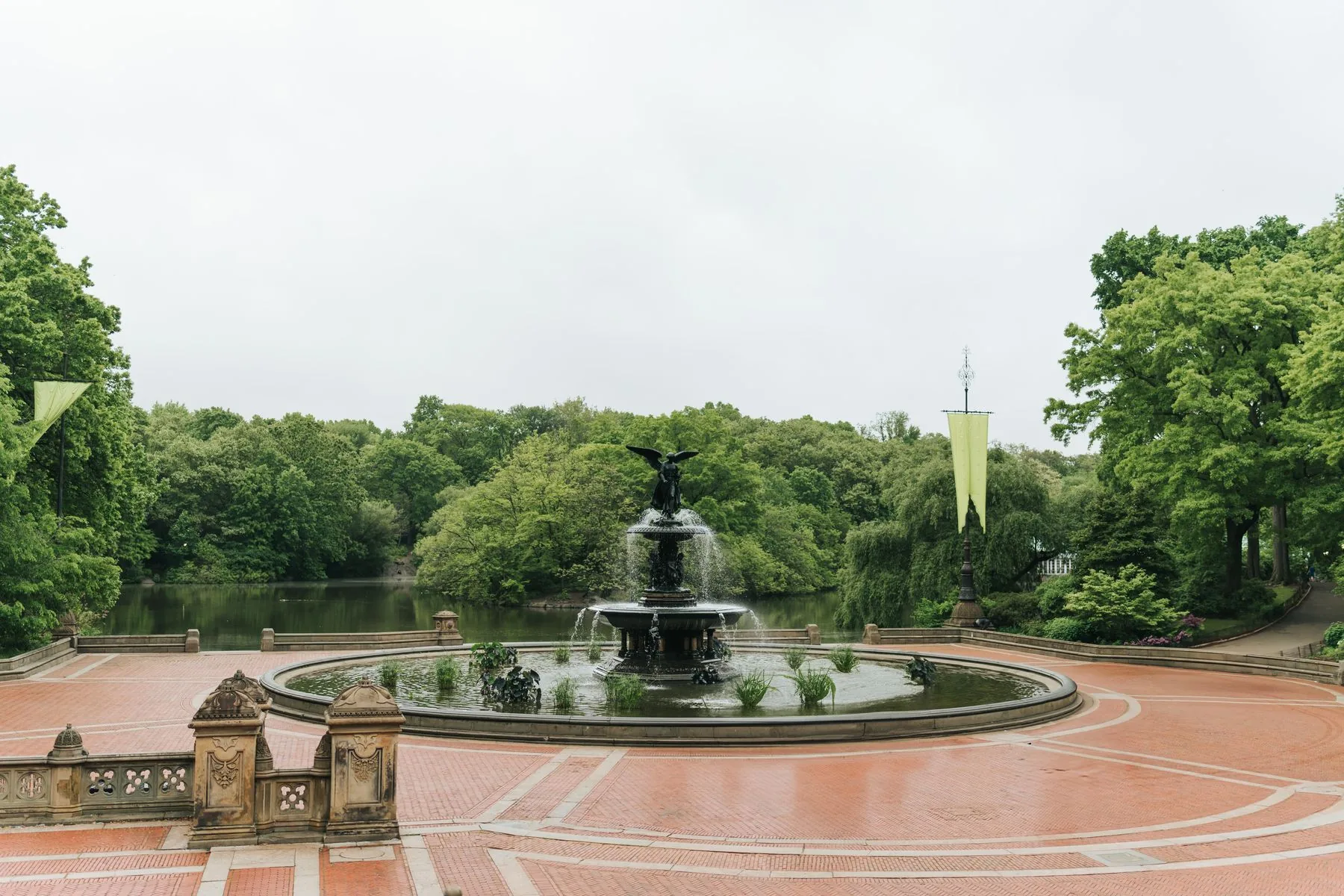 Bethesda Fountain’s winged Angel of the Waters statue above a circular pool, with brick terrace paving, the lake, and trees behind it.