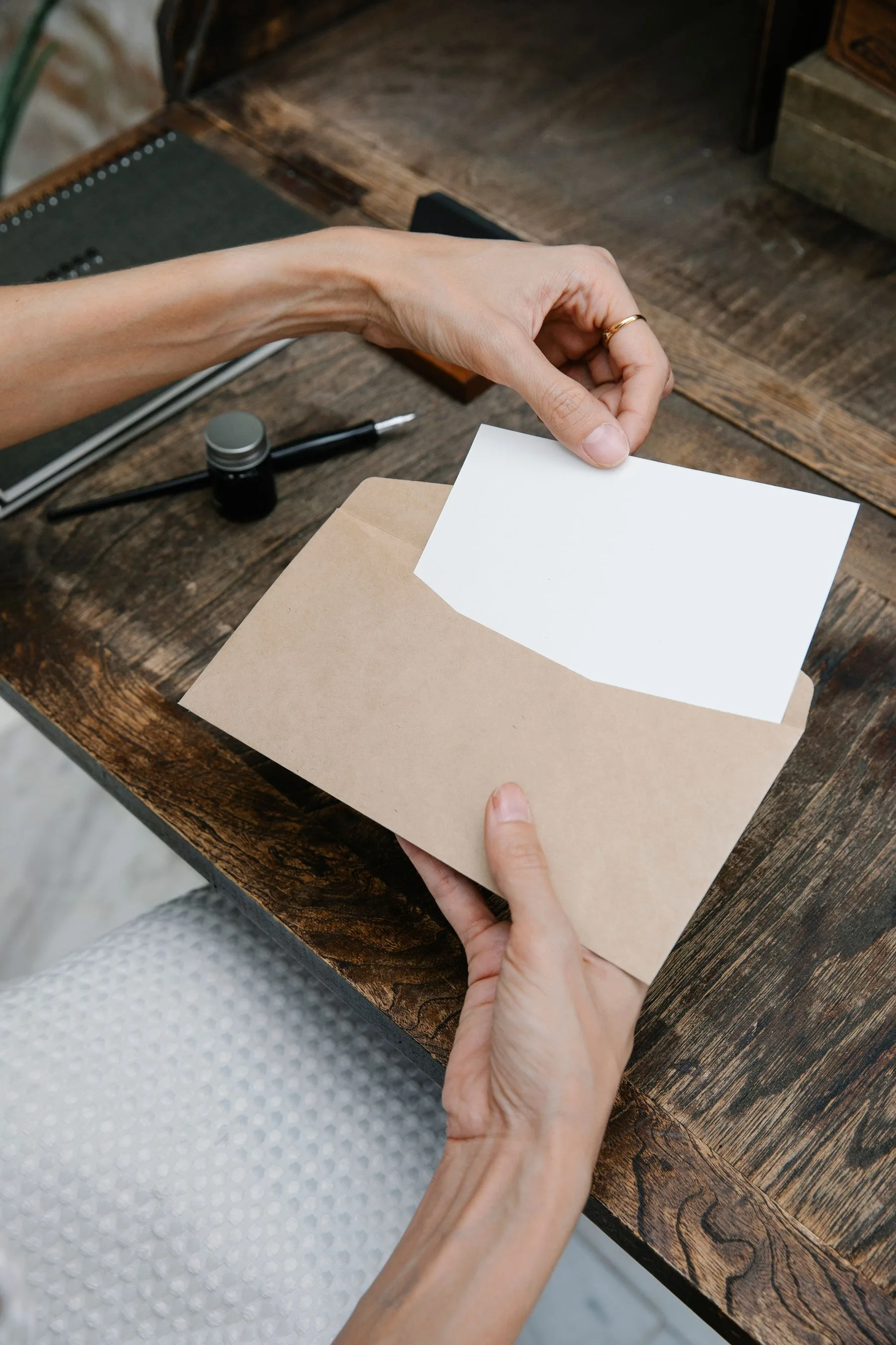 Hands holding a blank white card above a plain brown envelope on a wooden desk.
