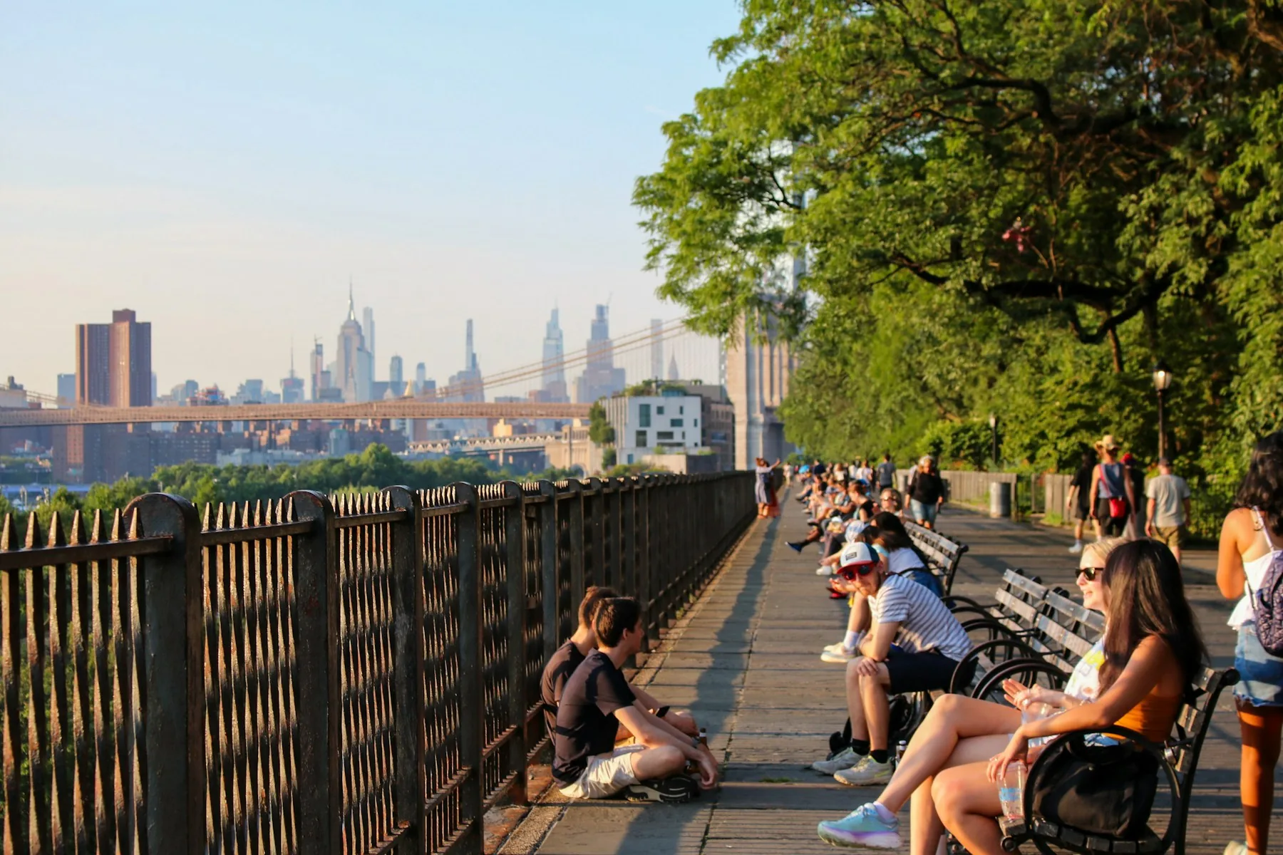 People sitting beside a tree-lined promenade in Brooklyn, with the Brooklyn Bridge and Manhattan skyline in the background.