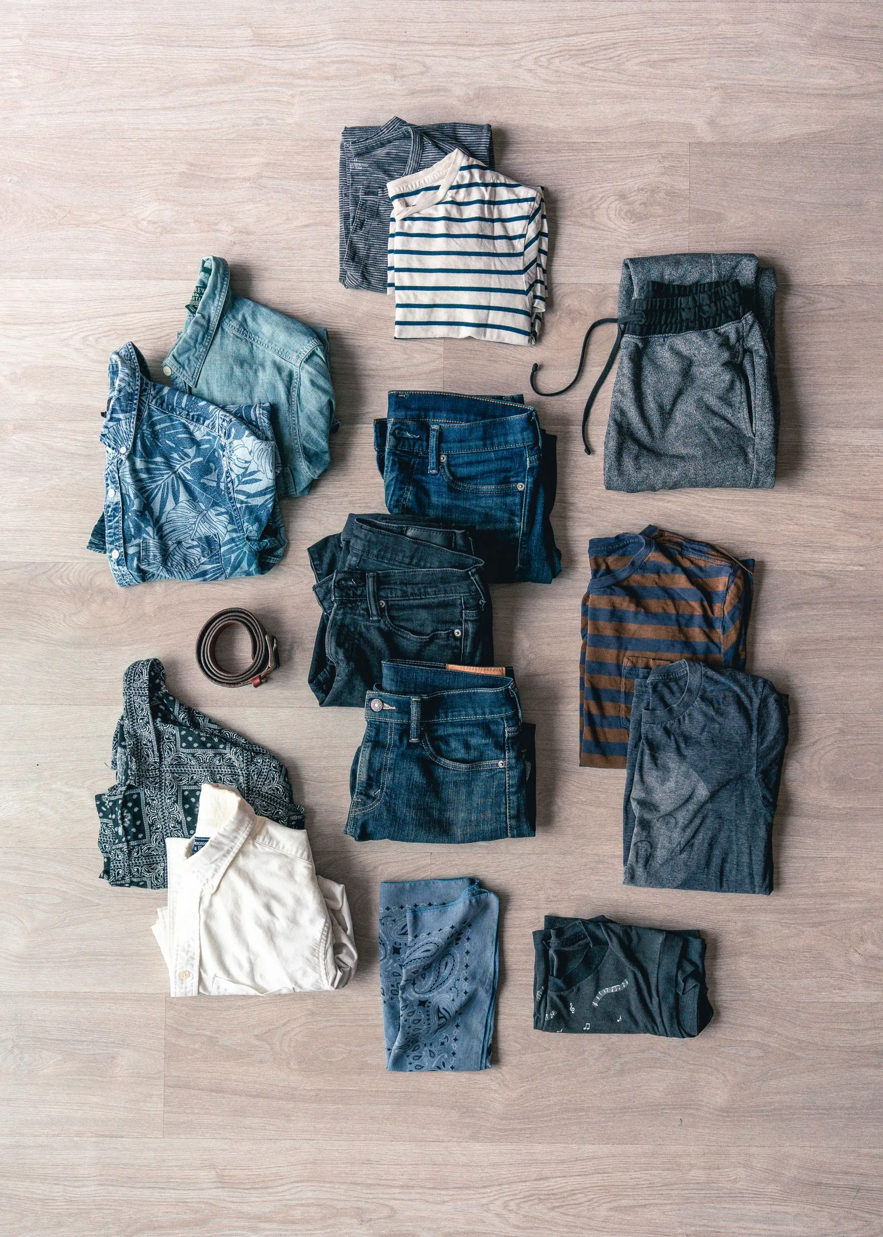 Folded casual shirts, T-shirts, jeans, and drawstring trousers arranged with a belt on a wooden floor.