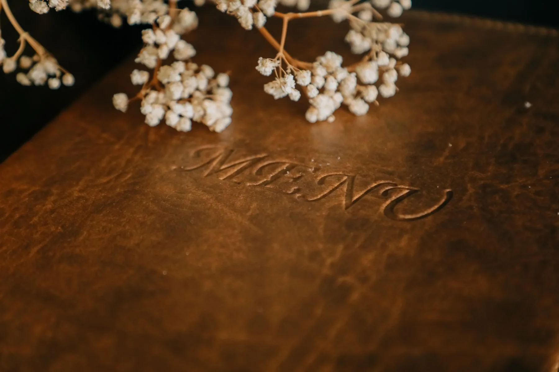 Close-up of a closed brown menu cover embossed “MENU” beneath dried flowers.