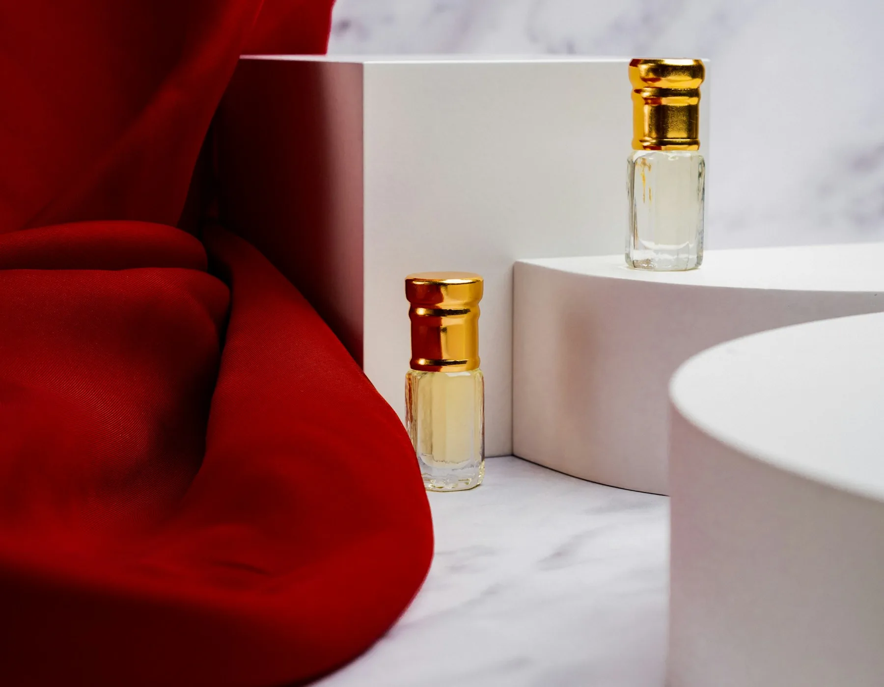 Two unbranded glass fragrance sample bottles with gold caps arranged on white blocks beside red fabric.