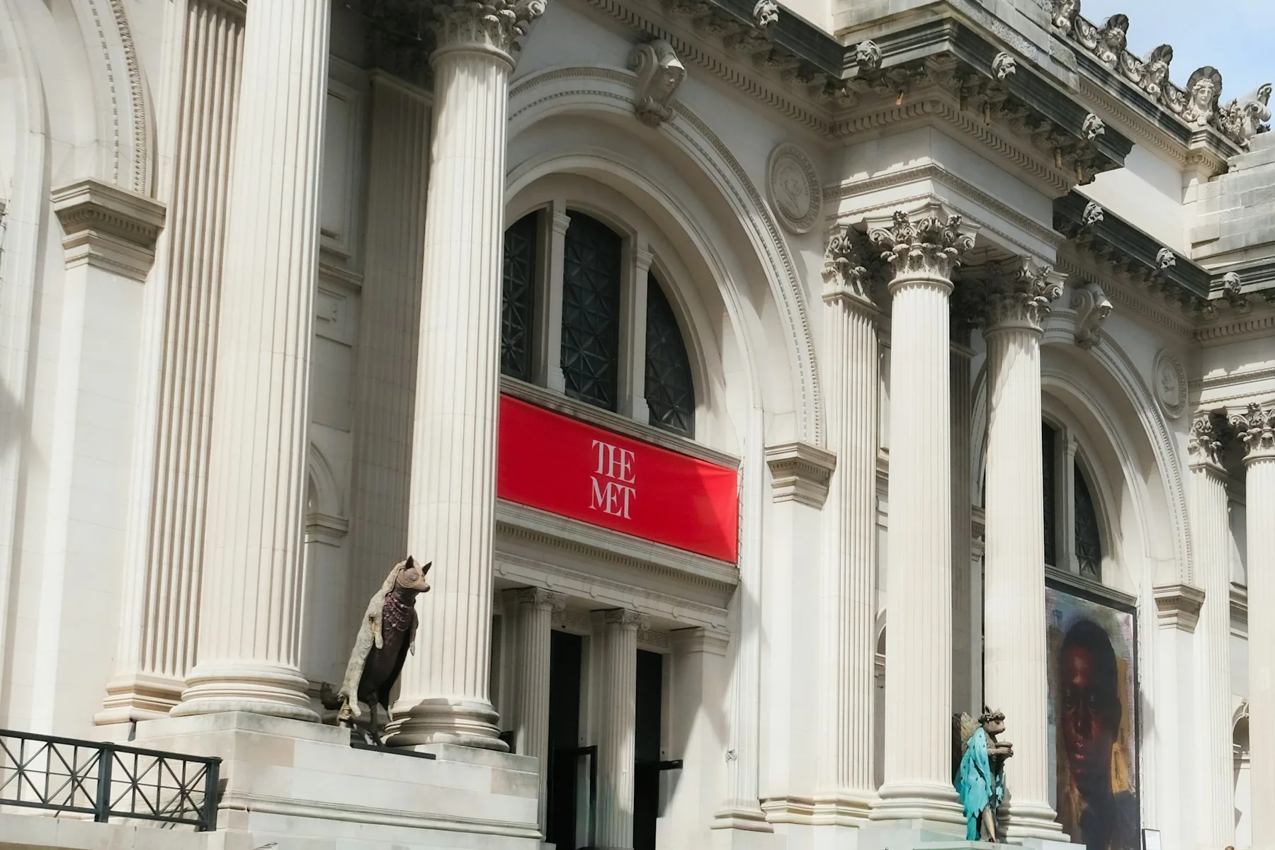 The Met Fifth Avenue entrance with tall stone columns, two statues and a red 'THE MET' banner.